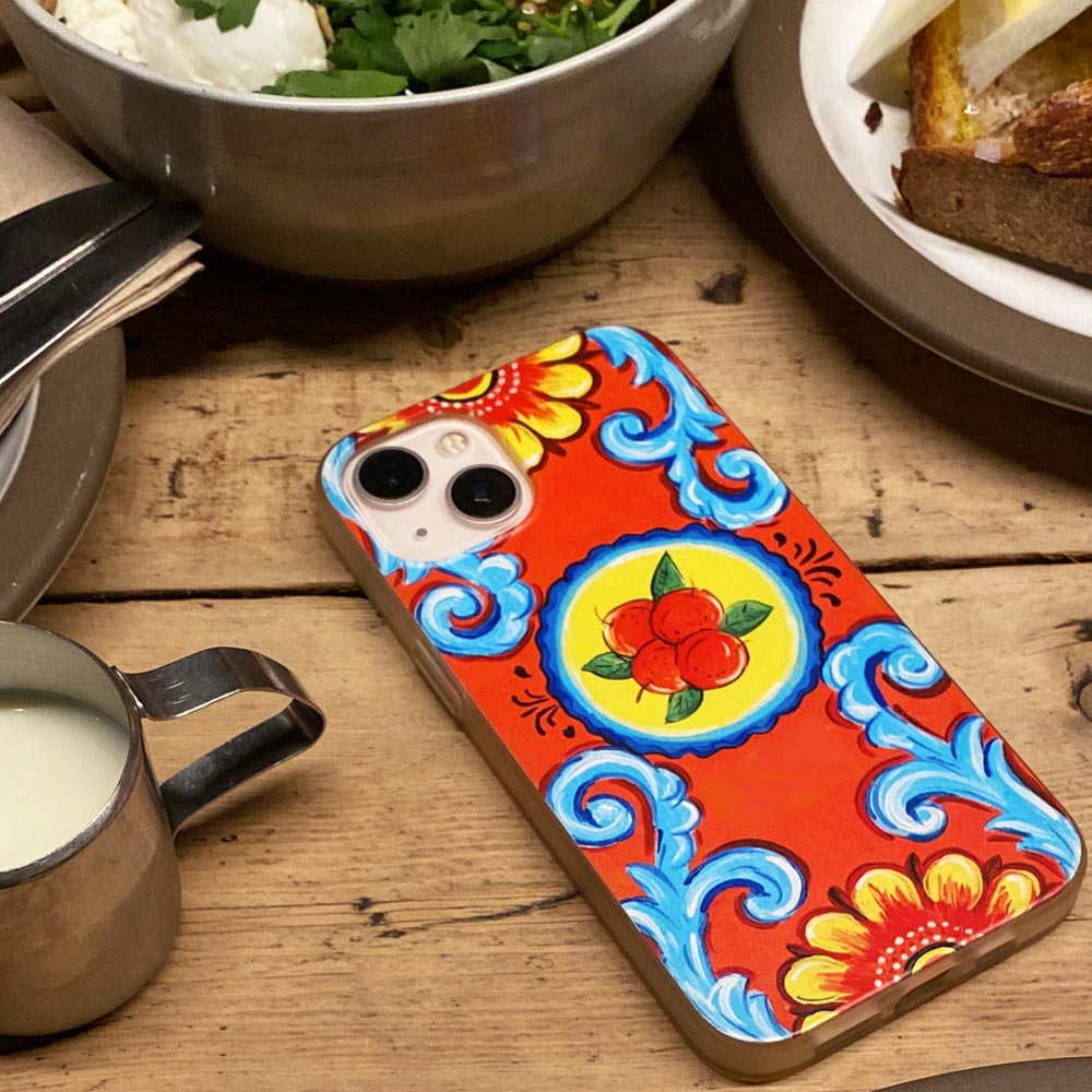 Colorful phone case with floral Sicilian design on a wooden table with food and a cup.