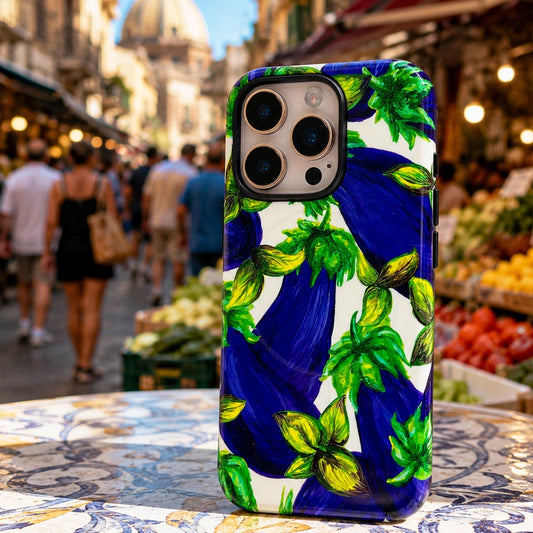 A phone case featuring a vibrant painted eggplant pattern in deep blue and green on a white background, shown standing on a tiled table with a blurred outdoor market scene in the background.