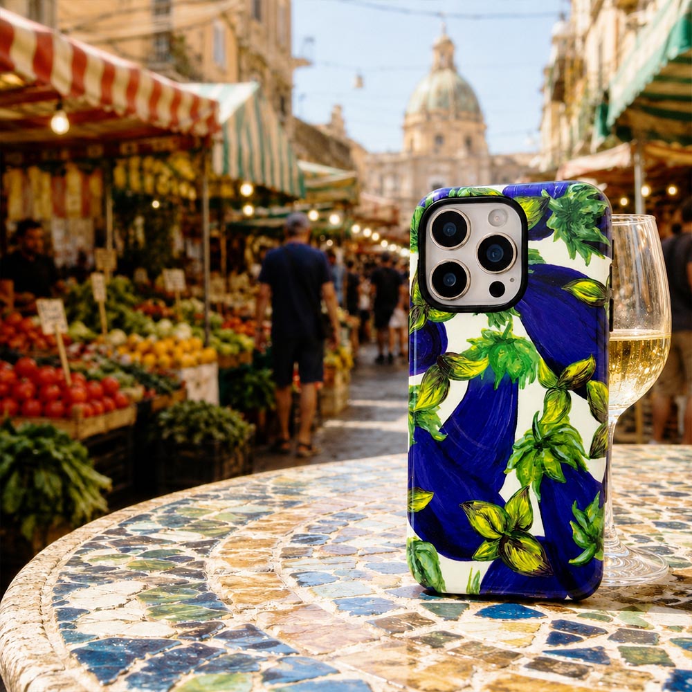 A phone case featuring a vibrant painted eggplant pattern in deep blue and green on a white background, shown standing on a tiled table with a blurred outdoor market scene in the background.
