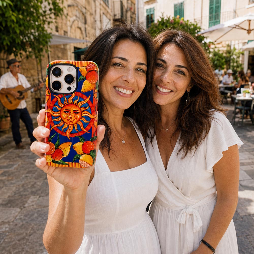 Two women taking a selfie outdoors in a sunny, historic town square. The woman on the left holds up a phone case featuring a vibrant, colorful sun and citrus fruit illustration on a dark blue background.